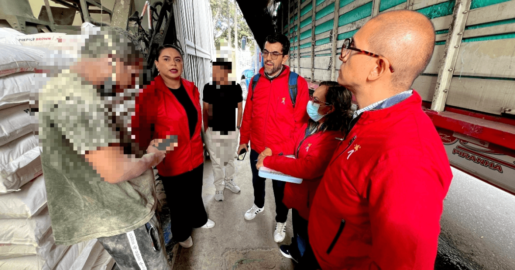 Personal con chaquetas rojas realiza una visita de inspección en una bodega de alimentos. Varias personas conversan entre pilas de sacos almacenados y un camión de carga estacionado junto al área de trabajo.