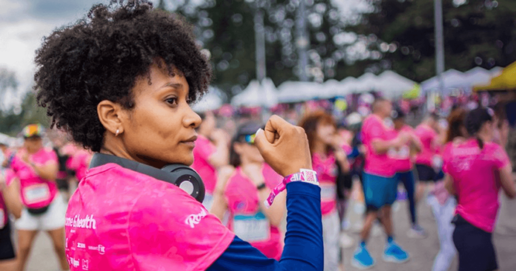 Participante de la Carrera de la Mujer con camiseta rosada y audífonos alrededor del cuello, entre corredoras reunidas en la zona de encuentro previa a la competencia.