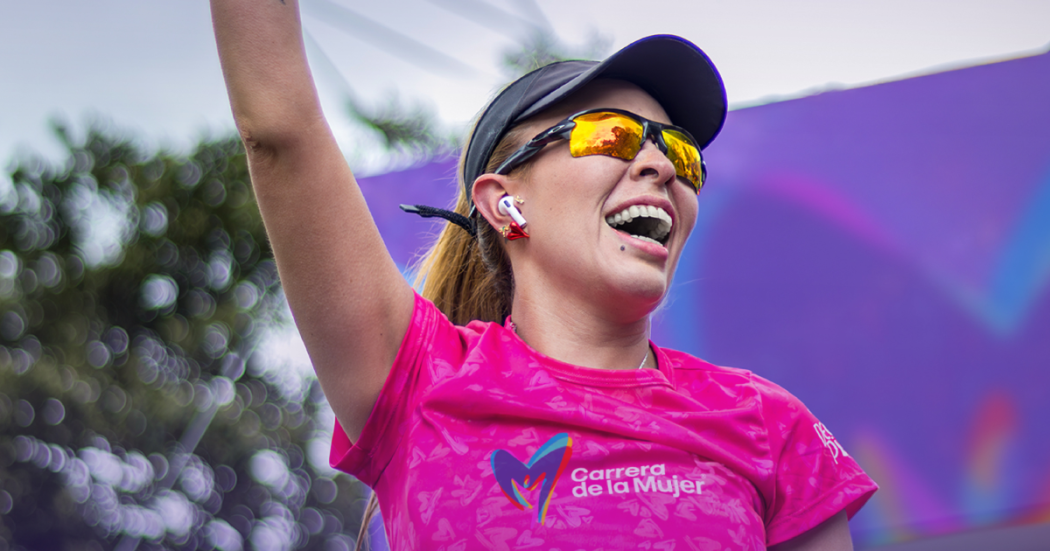 Participante de la Carrera de la Mujer con camiseta rosada, gorra negra, gafas deportivas y audífonos inalámbricos, levantando un brazo durante una actividad del evento al aire libre.