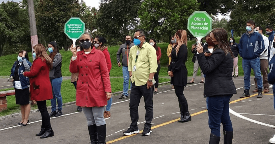 Grupo de personas participando en una actividad de evacuación al aire libre, posiblemente un simulacro de emergencia. Algunas llevan tapabocas y sostienen señales con los nombres de diferentes dependencias institucionales para el Simulacro Bogotá 2026.