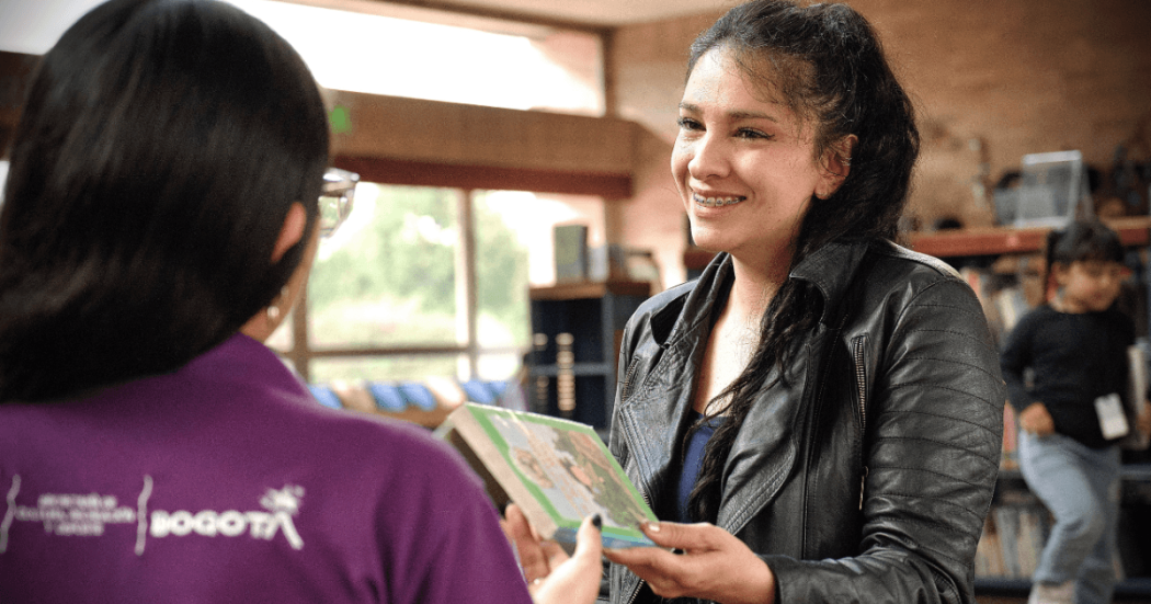 Imagen de una mujer sonriendo mientras entrega un libro a funcionaria de BibloRed de espaldas.