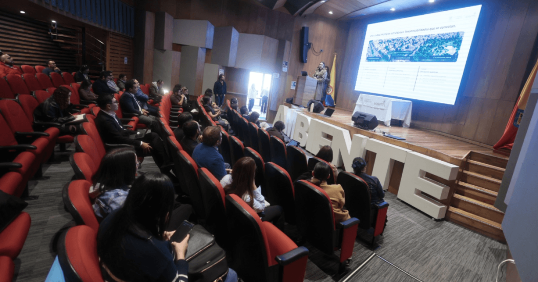 Asistentes participan en una jornada de socialización del Plan de Acción Acústico de Bogotá en un auditorio, mientras una presentación se proyecta en pantalla principal.