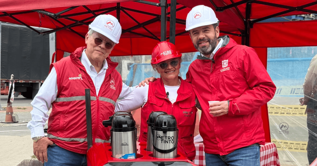 Alcalde Mayor de Bogotá, Carlos Fernando Galán, junto a Leonidas Narváez, gerente de la Línea 1 del Metro de Bogotá y Neida Ospina, vendedora de café en el carro ambulante 'Metro Tinto' visten con chalecos y cascos de seguridad rojos y blancos permanecen bajo una carpa roja junto a una mesa con termos de café. En las prendas se observan los logos de Bogotá y Metro de Bogotá.