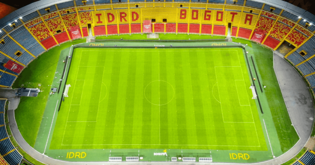 Vista general de la cancha del estadio El Campín con césped verde cortado en franjas longitudinales. En la parte superior se observan graderías con sillas amarillas y rojas, una franja publicitaria del IDRD y parte de la cubierta del escenario deportivo.
