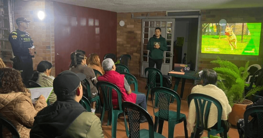Charla informativa sobre el cuidado de animales de compañía en un salón comunitario. Varias personas sentadas en sillas de plástico observan una presentación proyectada en pantalla, mientras una persona expone al frente y un agente de policía permanece de pie al lateral con proyección en pared de un perro en zona verde.
