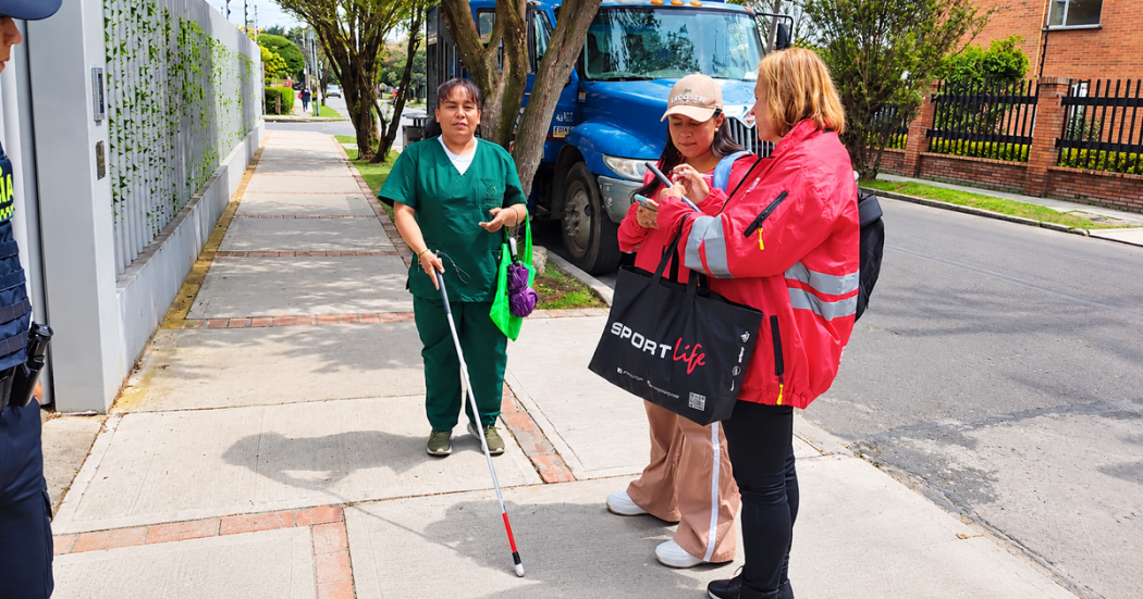 Foto de una mujer con discapacidad visual utilizando un bastón blanco con punta roja en una acera de Bogotá, mientras dos mujeres conversan a su lado.