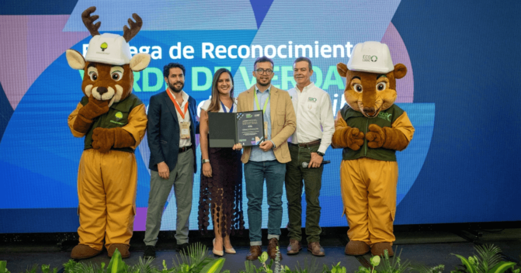 Entrega de reconocimiento ambiental a una entidad distrital. En una tarima, varias personas sostienen un galardón mientras posan para una fotografía, acompañadas por una mascota institucional. Al fondo se observa una pantalla azul con texto relacionado con el programa "Sello Verde de Verdad".