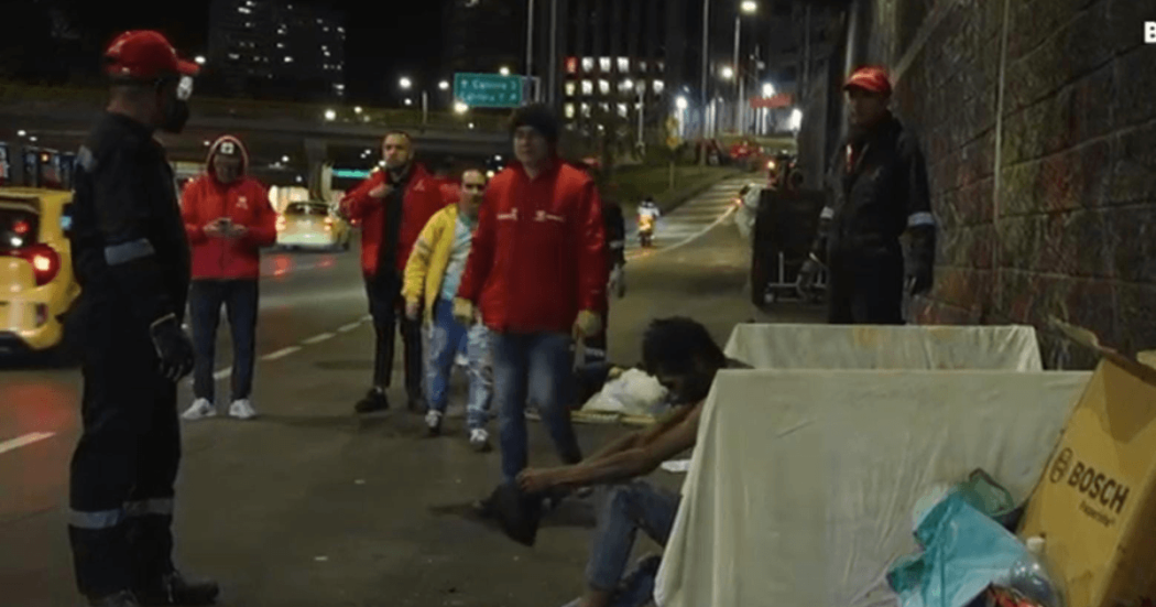 Operativo nocturno de atención social en un corredor vial, con personal de entidades distritales y varias personas junto a enseres y objetos retirados del espacio público.