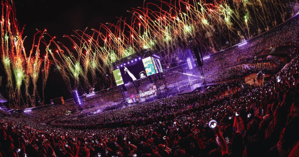 BTS en concierto en Bogotá con vista panorámica del estadio El Campín lleno de asistentes. En el escenario central hay pantallas gigantes y sobre el recinto se observan fuegos artificiales de colores que iluminan al público y las graderías.