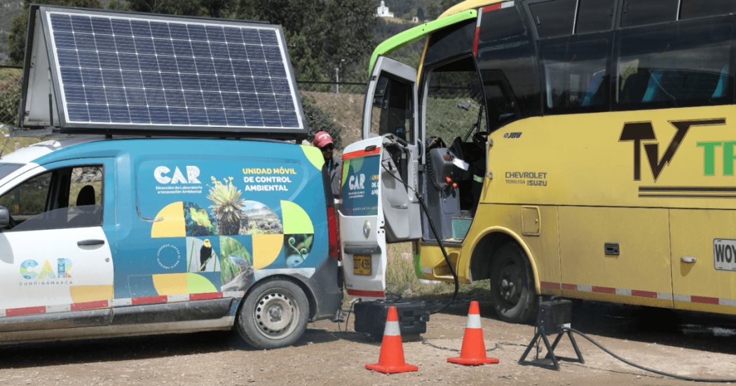 Vehículo de la Unidad Móvil de Control Ambiental de la CAR Cundinamarca, equipado con panel solar en el techo, conectado a un bus de transporte mediante cables de medición.