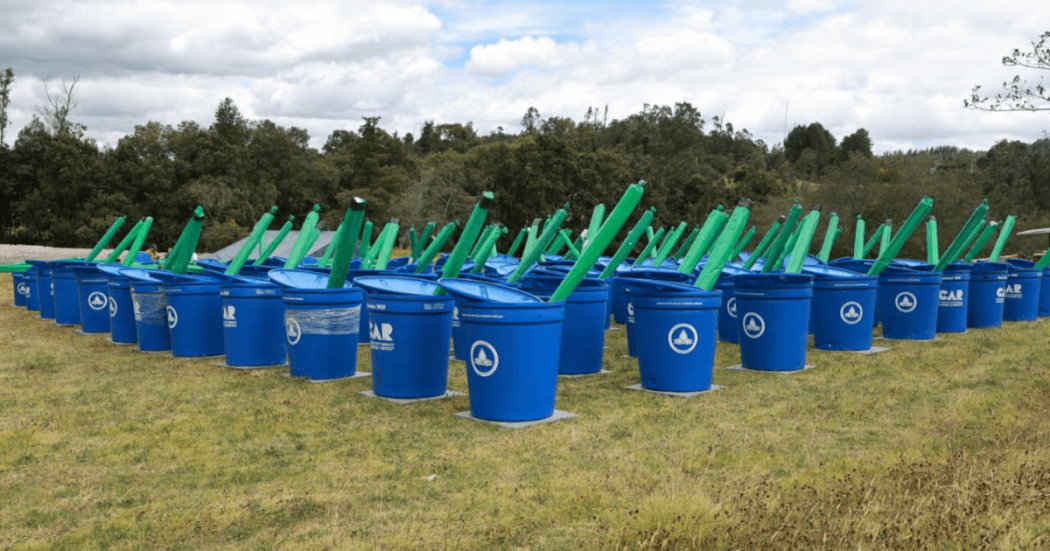 Canecas azules de la CAR Cundinamarca para almacenamiento de agua alineadas sobre un terreno cubierto de césped, junto a estructuras verdes instaladas como parte de un sistema de recolección y aprovechamiento de agua en zona rural.