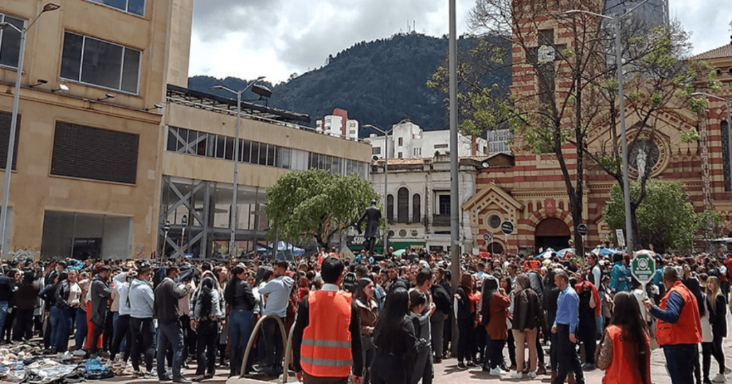 Personas reunidas en una plaza del centro de Bogotá durante una actividad al aire libre. En primer plano se observan participantes y personal con chalecos naranjas, mientras al fondo se distinguen edificaciones históricas y los cerros orientales.