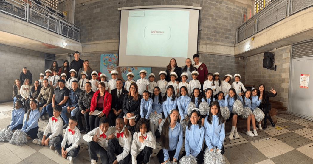 Grupo de estudiantes y docentes de Teusaquillo posa para una fotografía grupal en un espacio interior de un colegio. Al fondo se observa una pantalla de proyección y un muro de ladrillo, mientras los participantes están ubicados en varias filas frente a la cámara