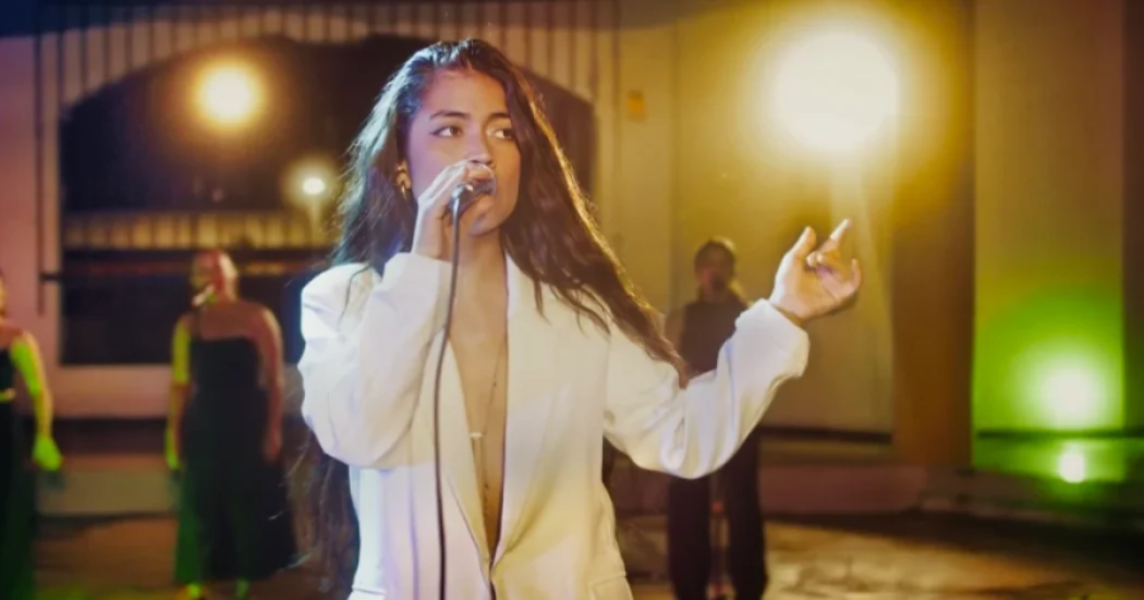 Imagen de una mujer joven de cabello rubio largo cantando con un micrófono.