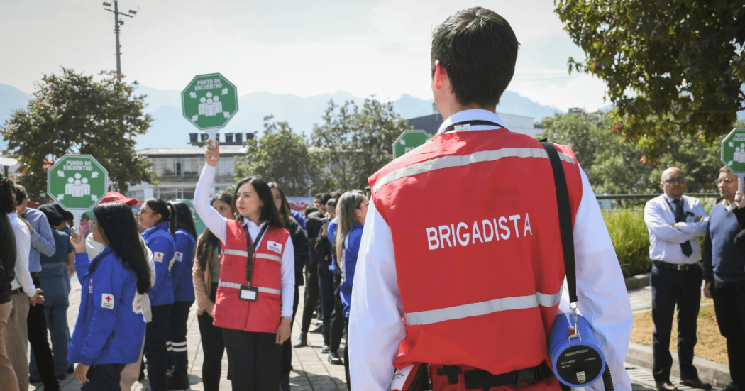 Brigadistas de la Cruz Roja Bogotá orientan un ejercicio de preparación para emergencias mientras varias personas sostienen señales de punto de encuentro en un espacio abierto.