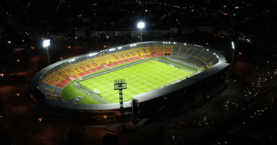 Vista nocturna del estadio El Campín iluminado durante un partido de fútbol, con las tribunas visibles alrededor del terreno de juego y las luminarias encendidas sobre la cancha.