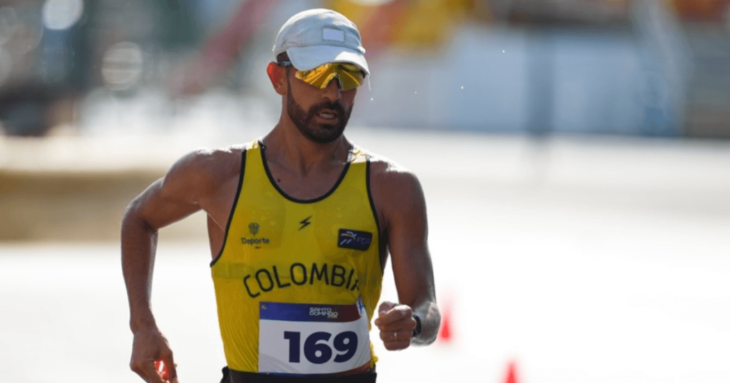 Eider Arévalo Deportista de marcha atlética con uniforme amarillo de la selección Colombia durante una competencia. Lleva gafas deportivas, gorra clara y un número de competencia visible en el pecho, mientras avanza por el circuito.