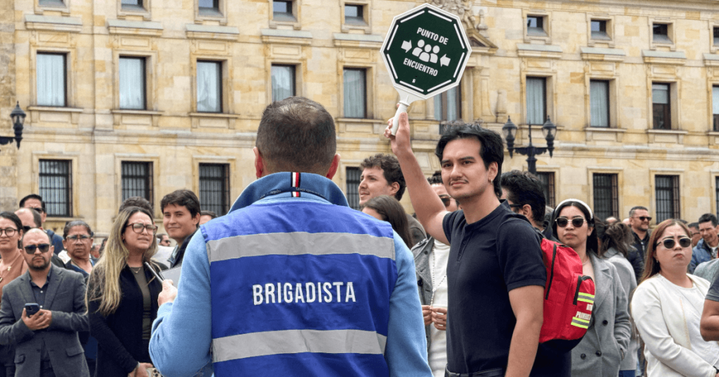 Grupo de personas reunidas en una plaza frente a un edificio institucional. En primer plano, una persona con chaleco azul que dice “BRIGADISTA” observa a otra que sostiene una señal octagonal con el texto “PUNTO DE ENCUENTRO”.