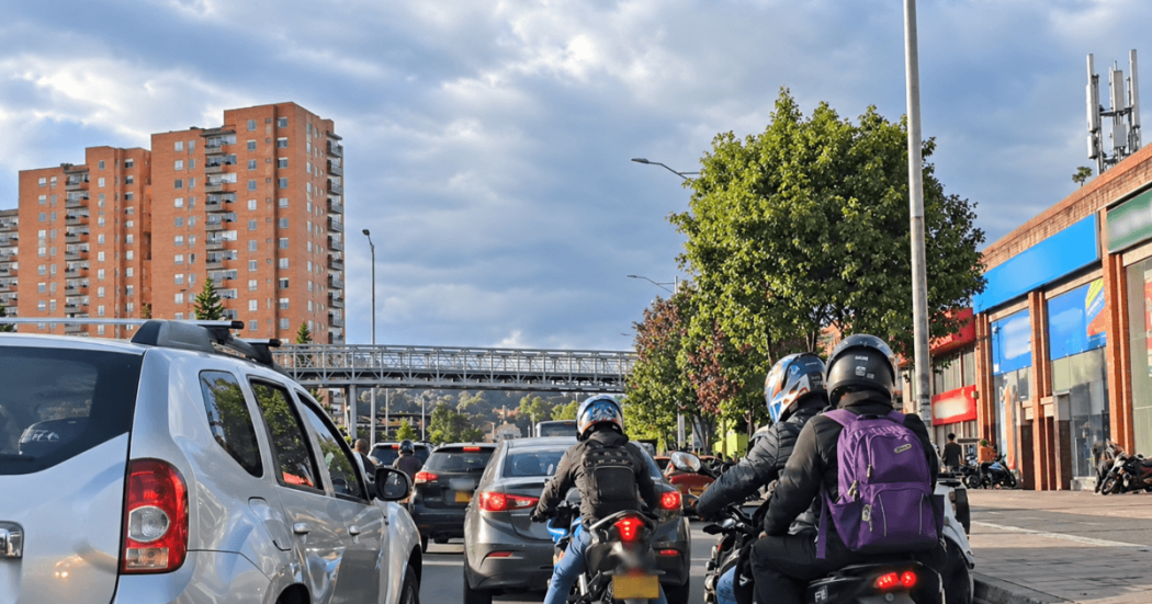 Motociclistas avanzan entre vehículos en una vía de Bogotá durante el día. A un costado de la carretera se observan árboles y, al fondo, edificios residenciales bajo un cielo parcialmente nublado.