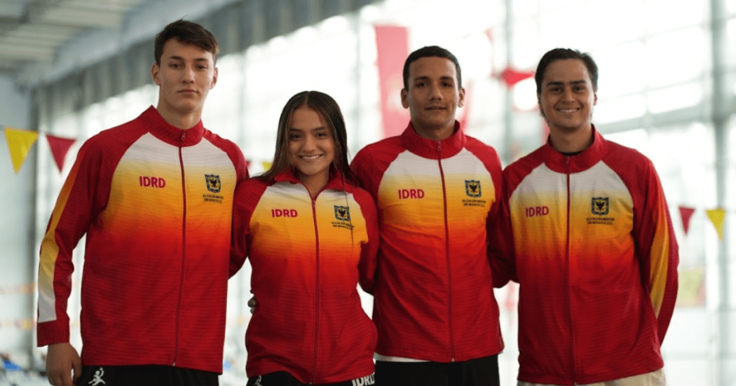 Cuatro deportistas del Equipo Bogotá posan juntos en una instalación deportiva cubierta. Llevan chaquetas de equipo con degradado rojo, naranja y amarillo, cremallera frontal y los textos visibles ‘IDRD’ y ‘Alcaldía Mayor de Bogotá D.C.’ en el pecho.
