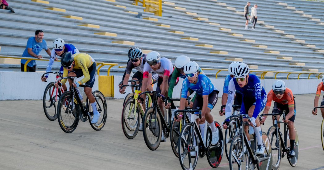 Grupo de ciclistas compite en una pista inclinada de velódromo durante una prueba de ciclismo. Las y los deportistas avanzan en fila sobre bicicletas de ruta, con uniformes de distintos colores y cascos de competición, mientras recorren la curva de la pista de madera.