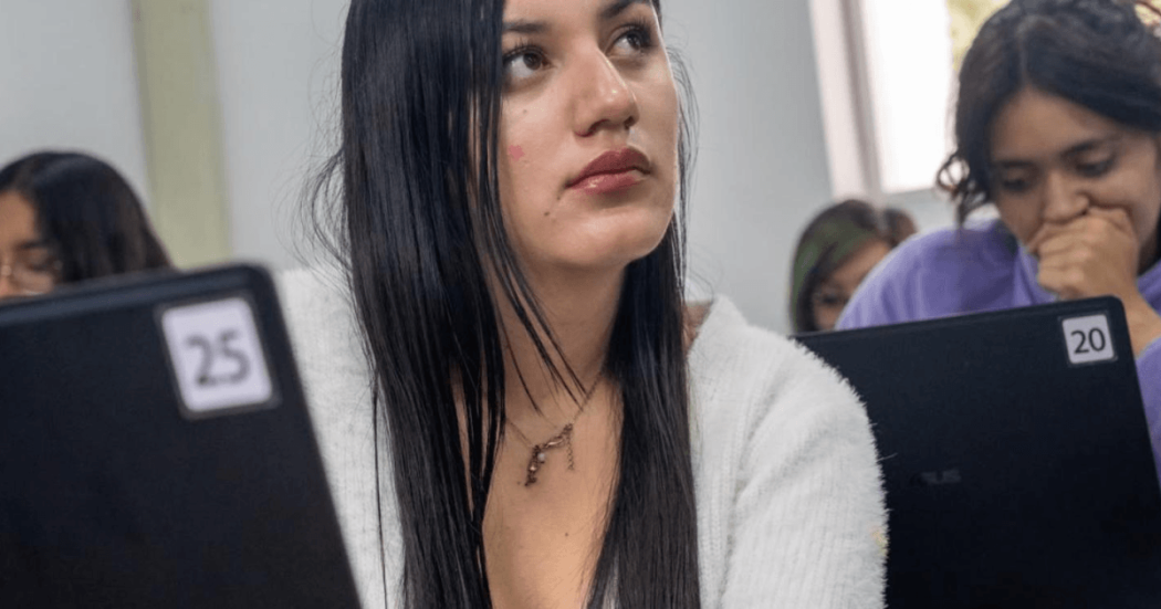 Foto de una mujer con otras dos de fondo, en un salón de clases.