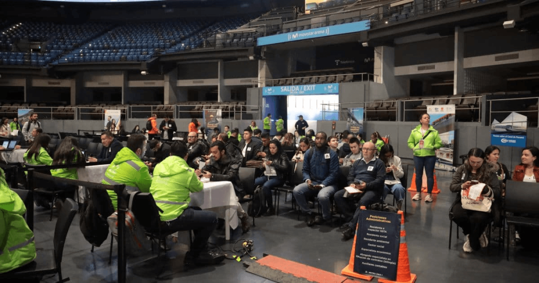 Personas sentadas en filas dentro de un recinto ferial mientras participan en una jornada de empleo. En primer plano hay mesas de atención y personas con chalecos institucionales; al fondo se observan estands, pantallas informativas y asistentes reunidos.