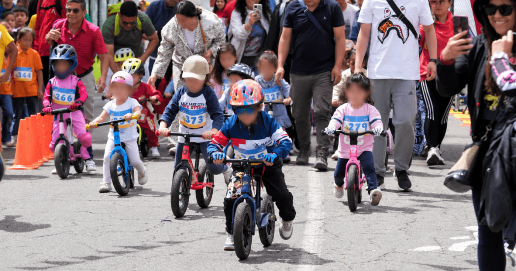 Niñas y niños participan en una carrera de bicicletas sin pedales en una vía urbana, acompañados por adultos y usando cascos de protección.