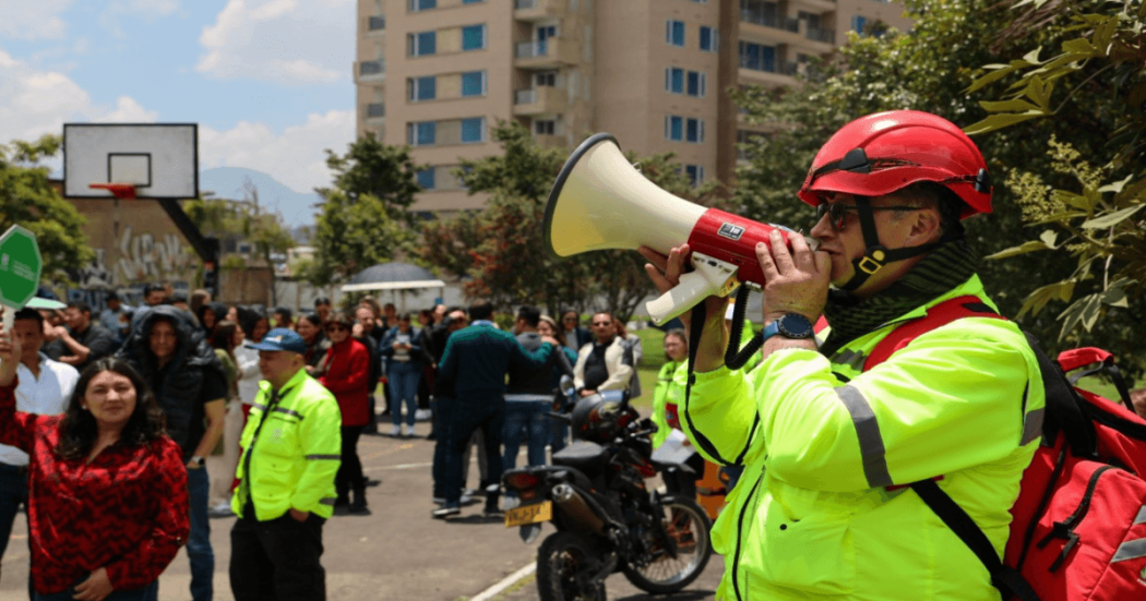 Persona con casco rojo y chaqueta reflectiva usa un megáfono para orientar a un grupo de ciudadanos durante un ejercicio de simulación de emergencia por sismo y otras emergencias en un espacio urbano de Bogotá.