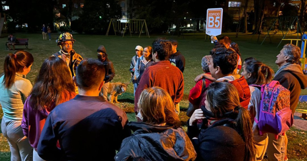 Grupo de personas reunidas en un parque durante una jornada nocturna de preparación para simulacro. Un integrante del cuerpo de bomberos brinda indicaciones mientras una persona sostiene un letrero con la identificación "Edificio B5".