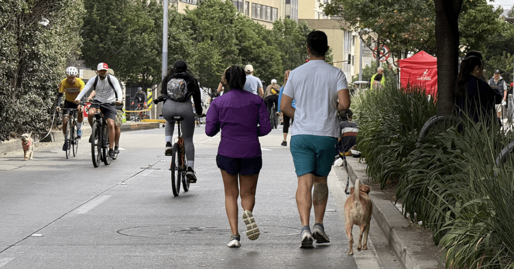 Personas caminando, corriendo y montando en bicicleta por una vía urbana cerrada al tráfico durante una jornada recreativa. Varias bicicletas circulan por la calzada y dos perros acompañan a los participantes en la Ciclovía de Bogotá.