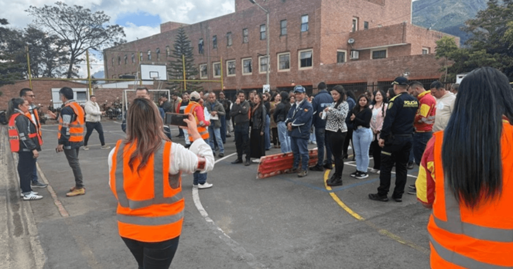 Grupo de personas reunidas en una cancha deportiva durante un ejercicio de simulacro en Bogotá. En primer plano, varias personas usan chalecos reflectivos naranjas mientras orientan a los participantes.