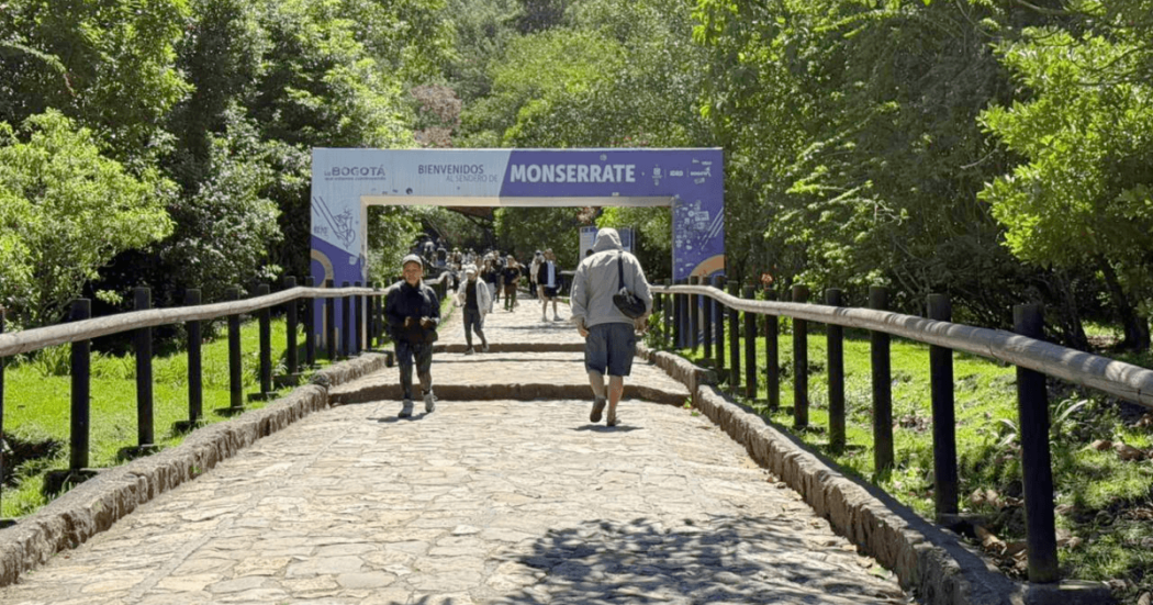 Personas caminan por el sendero peatonal de Monserrate mientras una persona se desplaza en bicicleta. Al fondo se observa un arco informativo de ingreso rodeado de vegetación y barandas de madera.