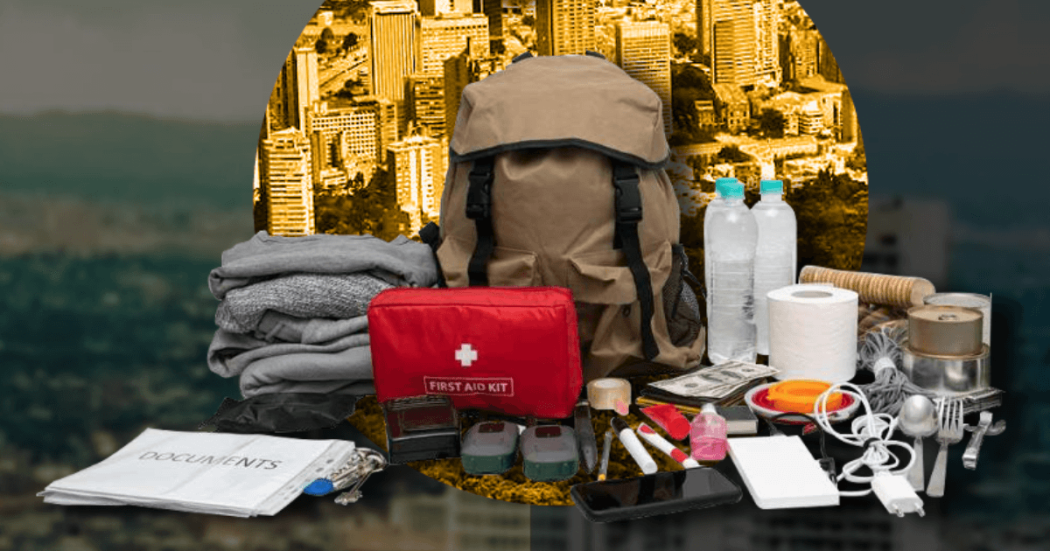 Kit de emergencias sobre una mesa con una mochila gris, botiquín rojo, linterna, pilas, radio portátil, alimentos no perecederos, botellas de agua y otros elementos recomendados para atender una emergencia.