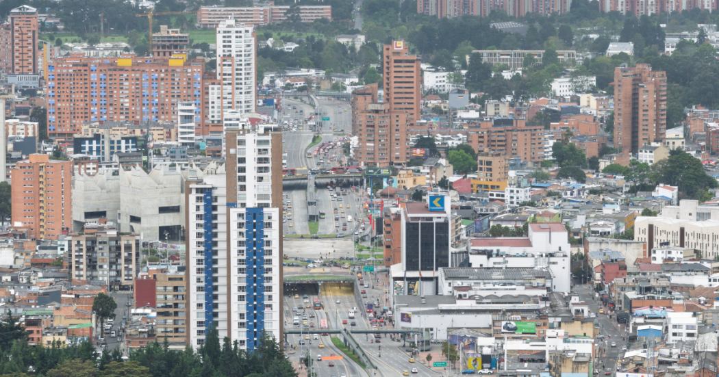 Vista panorámica de un sector urbano de Bogotá con edificios residenciales y comerciales de diferentes alturas, una vía principal con tráfico vehicular y construcciones densamente distribuidas en el paisaje urbano.