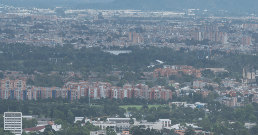 Vista panorámica de Bogotá bajo cielo nublado. En primer plano se observan edificios residenciales rodeados de árboles y zonas verdes, mientras al fondo se aprecia una amplia extensión urbana con edificaciones de diferentes alturas cubiertas por una ligera neblina.