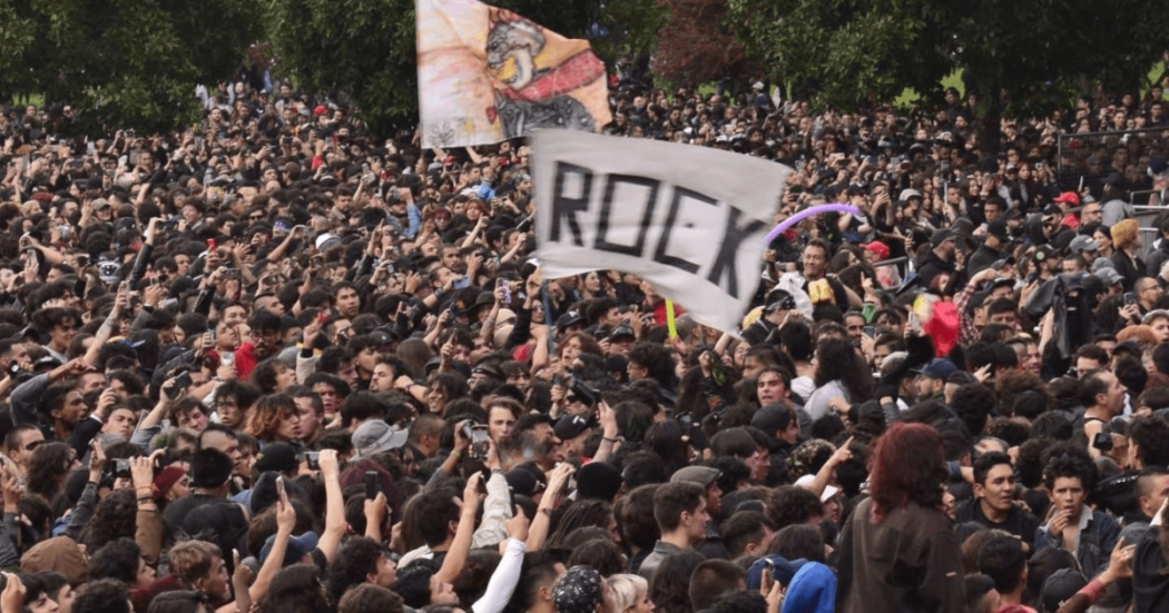 Multitud de asistentes reunidos al aire libre durante Rock al Parque en Bogotá. En el centro se observa una bandera blanca con la palabra “ROCK”, mientras cientos de personas ocupan gran parte del espacio frente al escenario, rodeadas por árboles.