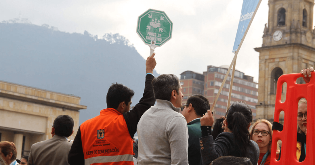 Personal de logística y participantes reunidos en una plaza del centro de Bogotá durante un simulacro. Una persona sostiene en alto una señal verde de “Punto de encuentro”, mientras alrededor se observan varios asistentes. Imagen de referencia para el Simulacro Bogotá 2026.