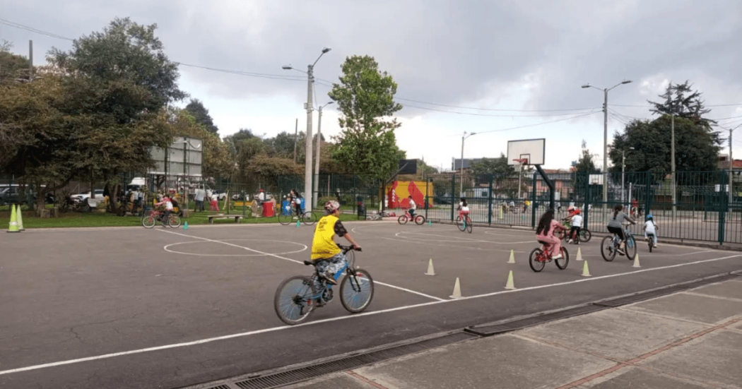 Persona con casco amarillo monta una bicicleta entre conos de señalización instalados en una pista durante Vacaciones Recreativas del IDRD con actividades de formación ciclista al aire libre.