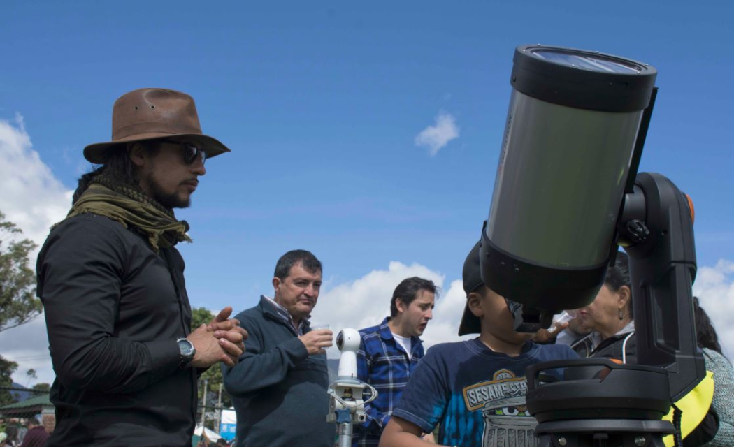 Un hombre enfrente de un telescopio, detrás hay varias personas en fila