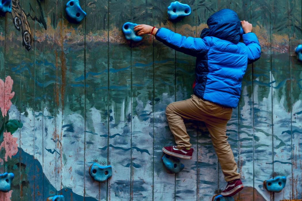 Un niño con chaqueta azul y pantalón café esta subiendo una pared de escalada 
