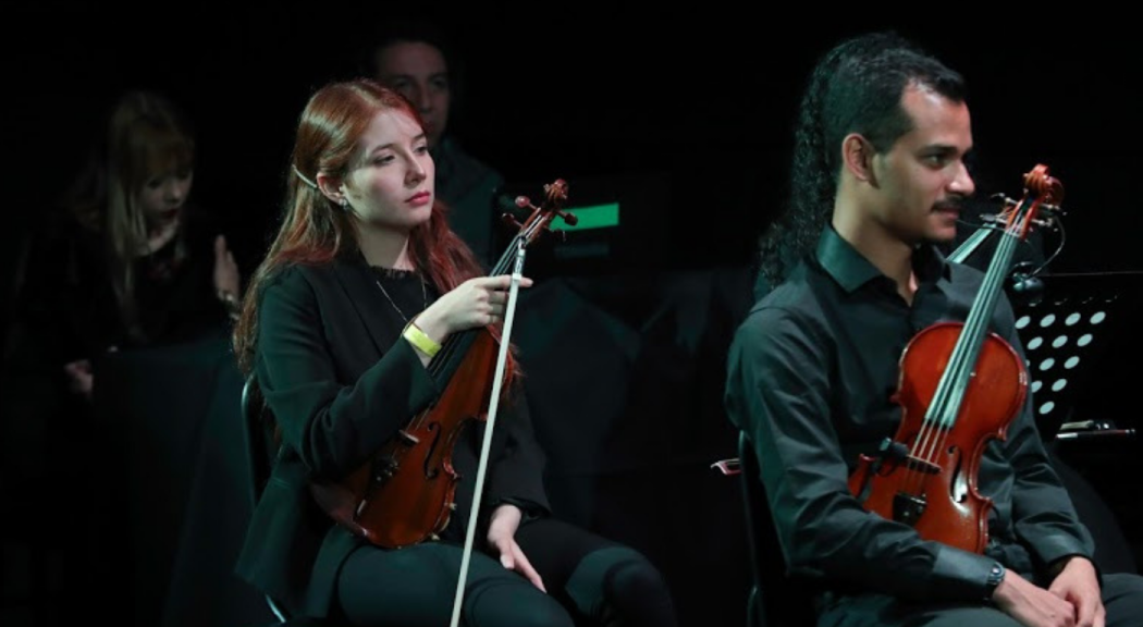 Una mujer y un hombre de la Orquesta Filarmónica Juvenil de Cámara sostienen sus instrumentos de cuerda en medio de una presentación.