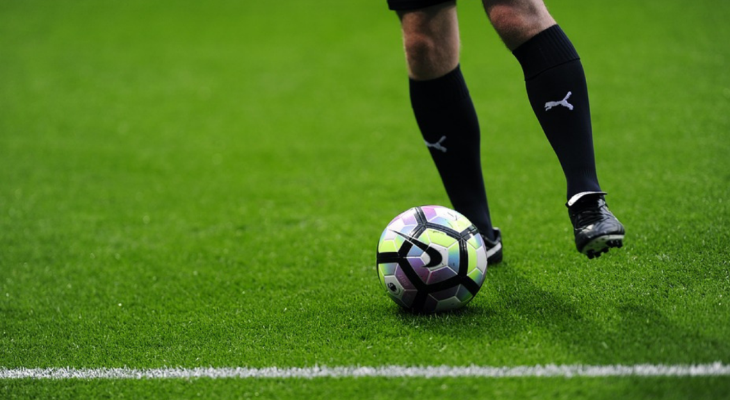 Foto de archivo. Primer plano de unas piernas con medias negra altas y guayos que patean un balón de fútbol negro con plano sobre el pasto verde oscuro.
