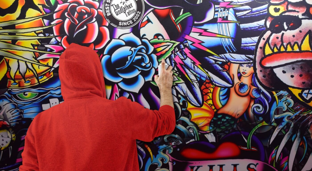 Un hombre usando saco con capota rojo hace un graffiti en una pared.
