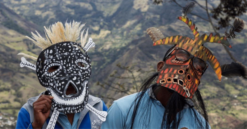 Imagen de dos hombres disfrasados luciendo sus máscaras, al fondo se ve paisaje y montañas