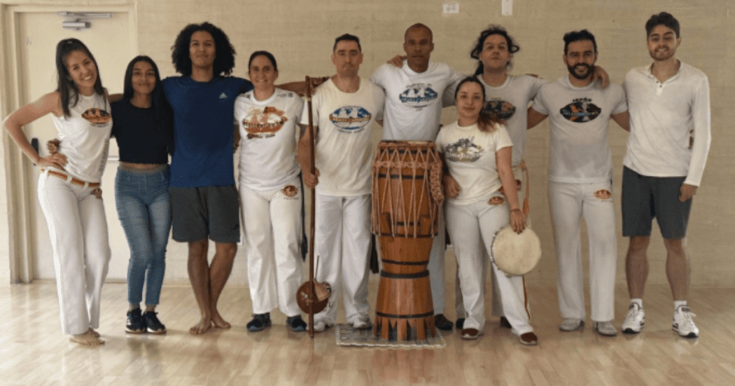 Imagen de varias personas adultas vestidas para practicar capoeira junto a instrumentos musicales propios de este arte