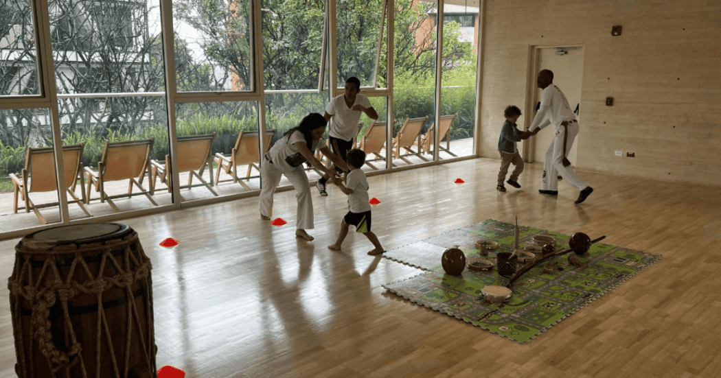 Imagen de niños en salón de baile siendo instruidos por adultos en capoeira