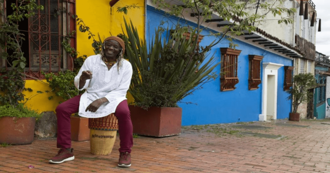 Imagen del maestro Nicoyembe sentado a las afueras de una casa antigua de color azul rey y amarillo