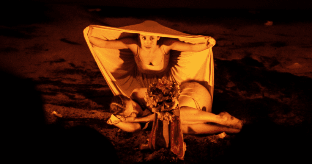 Imagen en fondo oscuro de dos mujeres, una acostada y otra de rodillas con un muñeco que representa la muerte