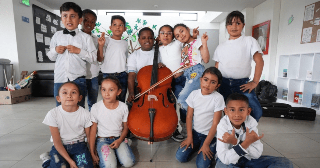 Foto de un grupo de niños y niñas posa sonriente alrededor de un violonchelo en un espacio escolar.